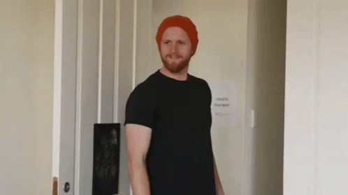Man In Red Hat And Black Shirt In Front Of Toilets This Way Sign GIF