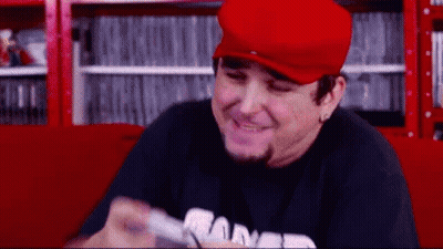 Man In Red Hat And Black Shirt Smiling Holding Remote GIF