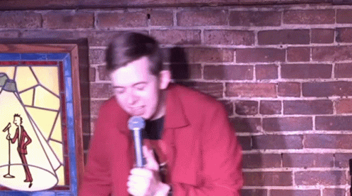 Man In Red Jacket Holding Microphone In Front Of Brick Wall GIF