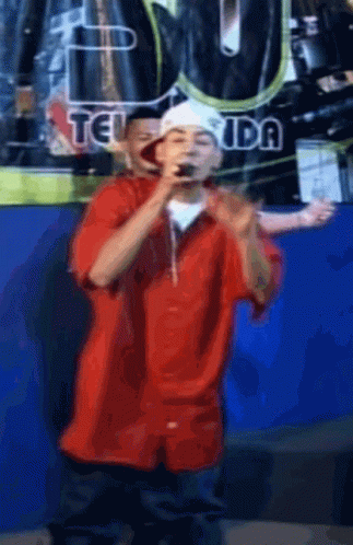 Man In Red Shirt Singing Into Microphone In Front Of Teiuda Sign GIF