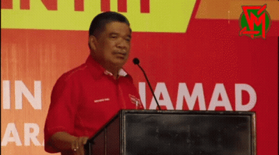 Man In Red Shirt Speaking At Podium In Jamad GIF