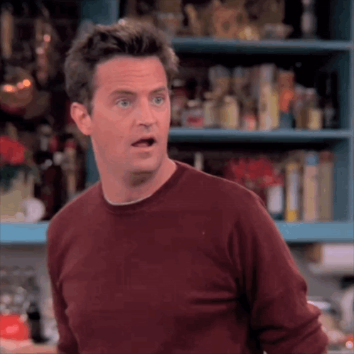 Man In Red Sweater Standing In Front Of Shelf Making Funny Face GIF