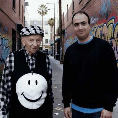 Man In Smiley Face Vest Beside Older Man GIF
