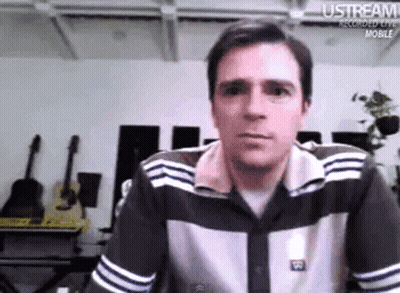 Man In Striped Shirt Sitting At Table Looking At Camera GIF
