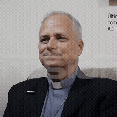 Man In Suit And Clergy Collar Smiling In Front Of Una Com Abri Sign GIF