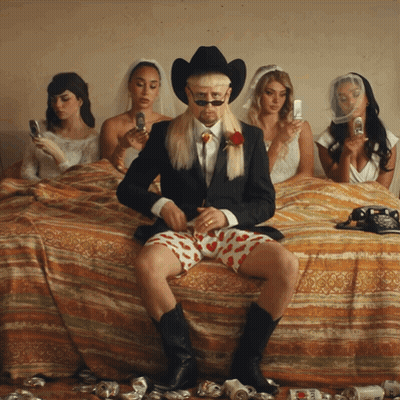 Man In Suit And Cowboy Hat On Bed Surrounded By Women GIF