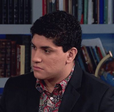 Man In Suit And Paisley Shirt Sitting Before Bookshelf GIF
