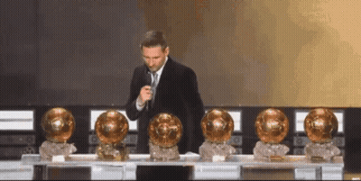 Man In Suit And Tie At Microphone With Gold Trophies GIF