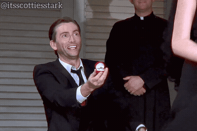 Man In Suit And Tie Holding Engagement Ring To Woman GIF