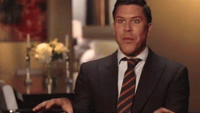Man In Suit And Tie Sitting Beside Vase Of Flowers GIF