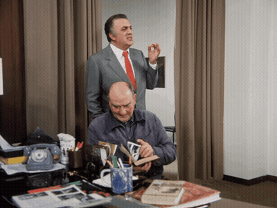Man In Suit Behind Man At Desk With Book GIF