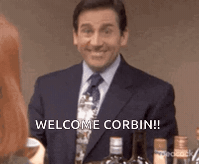 Man In Suit Saying Welcome Corbin Next To Woman GIF