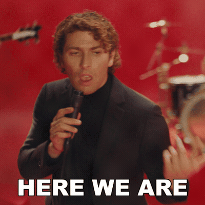 Man in Suit Singing Into Microphone Saying Here We Are