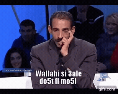 Man In Suit Sitting At Table With Hand On Chin And Crowd Behind GIF