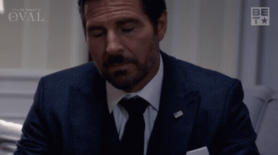 Man In Suit Sitting Before Oval Sign GIF
