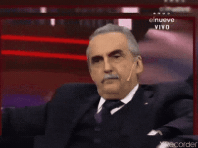 Man In Suit Sitting Before Screen Saying El Nueve Vivo GIF