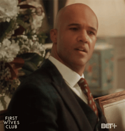 Man In Suit Sitting In Front Of First Wives Club Sign GIF