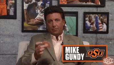 Man In Suit Sitting In Front Of Mike Gundy Sign GIF
