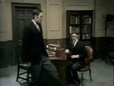 Man In Suit Standing Beside Man In Chair GIF