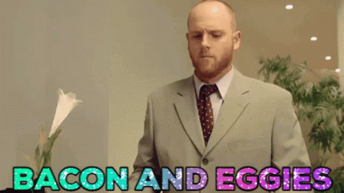 Man In Suit Standing In Front Of Sign Saying Bacon And Eggies GIF