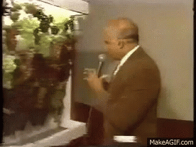 Man in Suit Talking Into Microphone in Front of Refrigerator Filled With Vegetables