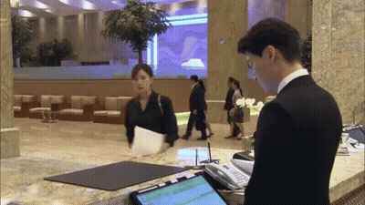 Man In Suit Talking To Woman At Counter In Lobby GIF