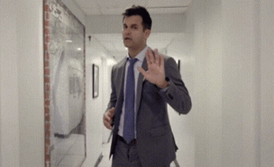 Man In Suit Walking Down Hallway With Hand Up GIF