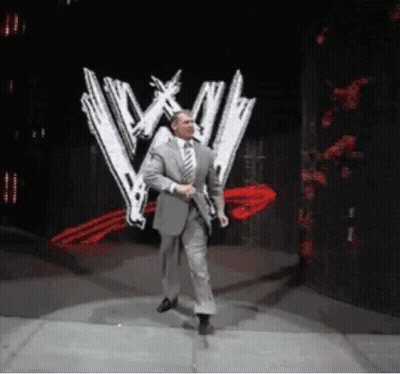 Man In Suit Walking On Stage In Front Of Wrestling Logo GIF