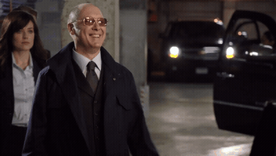 Man In Sunglasses And Suit Smiles With Woman Walking Behind GIF