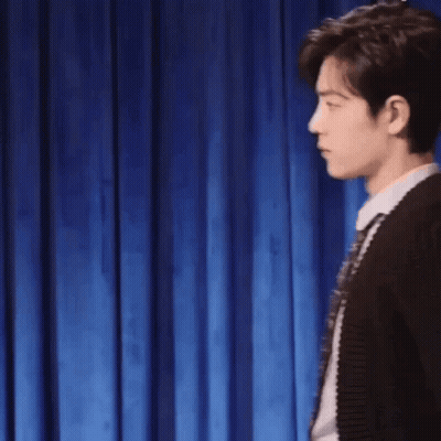 Man In Sweater And Tie Making Fist In Front Of Blue Curtain GIF