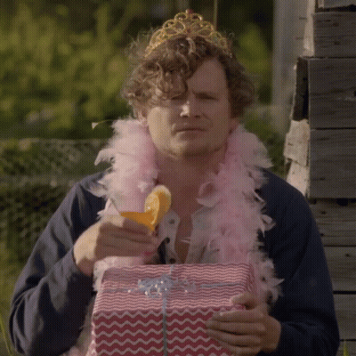 Man In Tiara And Feather Boa Holding Gift Box GIF