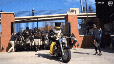 Man In Top Hat Riding Motorcycle Past Football Players GIF