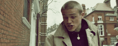 Man In Trench Coat Walking Down Street Next To Brick Building GIF