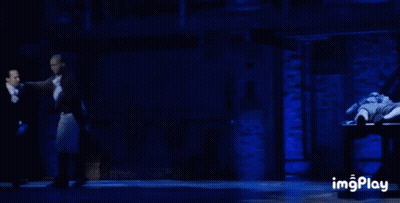Man In Tuxedo Dancing On Stage In Front Of Man Laying On Table GIF