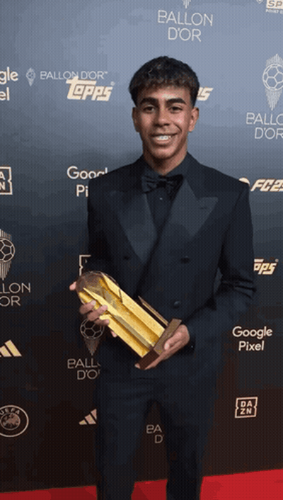 Man In Tuxedo Holding Gold Trophy In Front Of Ballon D'Or Wall GIF