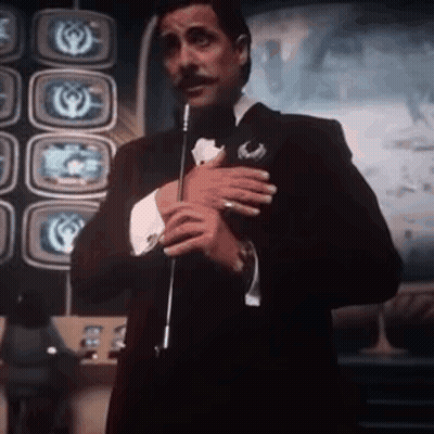 Man In Tuxedo Holding Microphone In Front Of Televisions GIF