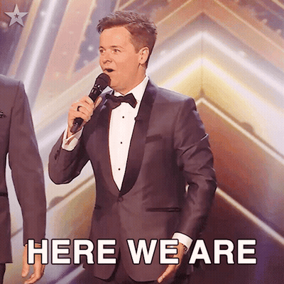 Man In Tuxedo Singing Into Microphone Here We Are GIF