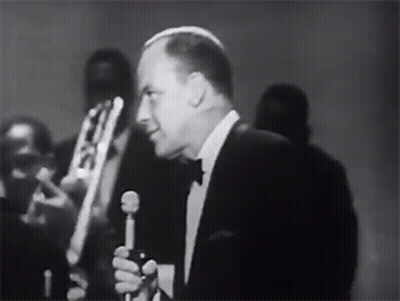 Man In Tuxedo Singing Into Microphone With Band Behind GIF