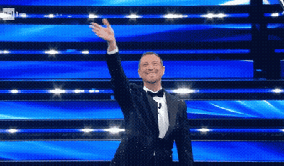 Man In Tuxedo Standing On Stage With Hand Up GIF