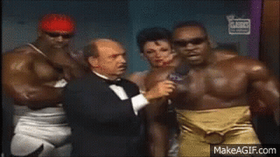 Man In Tuxedo Talking Into Microphone Beside Wrestlers GIF