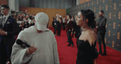 Man In White Hat Holding Microphone Next To Woman In Black Dress GIF