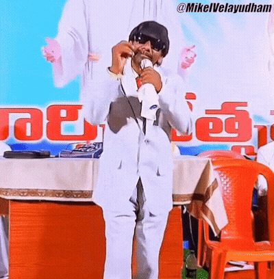 Man In White Suit Holding Microphone Before Mikelvelayudham Sign GIF
