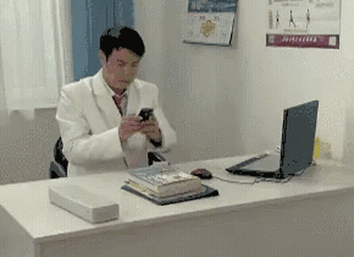 Man In White Suit Sitting At Desk Looking At Phone GIF