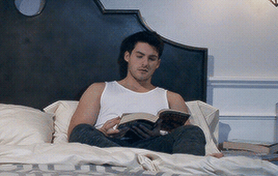 Man In White Tank Top Reading Book On Bed GIF