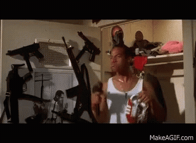 Man In White Tank Top Standing Surrounded By Guns GIF