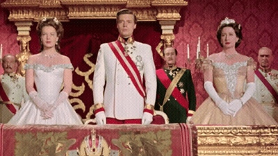 Man In White Uniform With Red And Gold Sash Between Two Women In White Dresses GIF