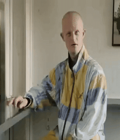 Man In Yellow And Blue Jacket Leaning Against Wall GIF