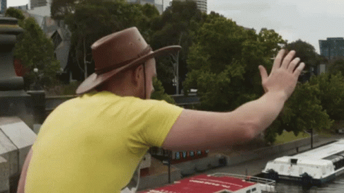 Man In Yellow Shirt And Brown Hat Waving At Boat GIF