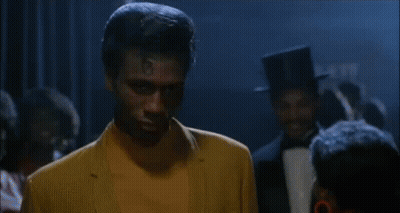 Man In Yellow Suit Standing With Group In Dark Room GIF