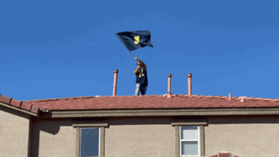 Man On Roof Holding Flag With Letter M GIF